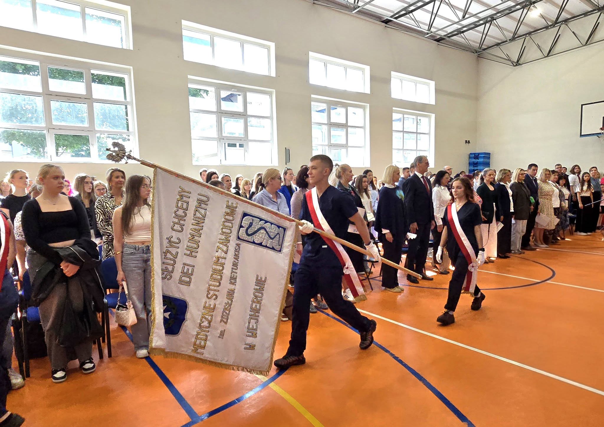 Zdjęcie grupowe nauczycieli i uczniów wraz z pocztem sztandarowym na Hali Sportowej.